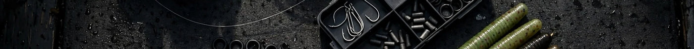 Top-down flat-lay of deadstick tackle on a dark boat deck: spinning rod, fluorocarbon line, wacky hooks, nail weights and soft plastic stick worms