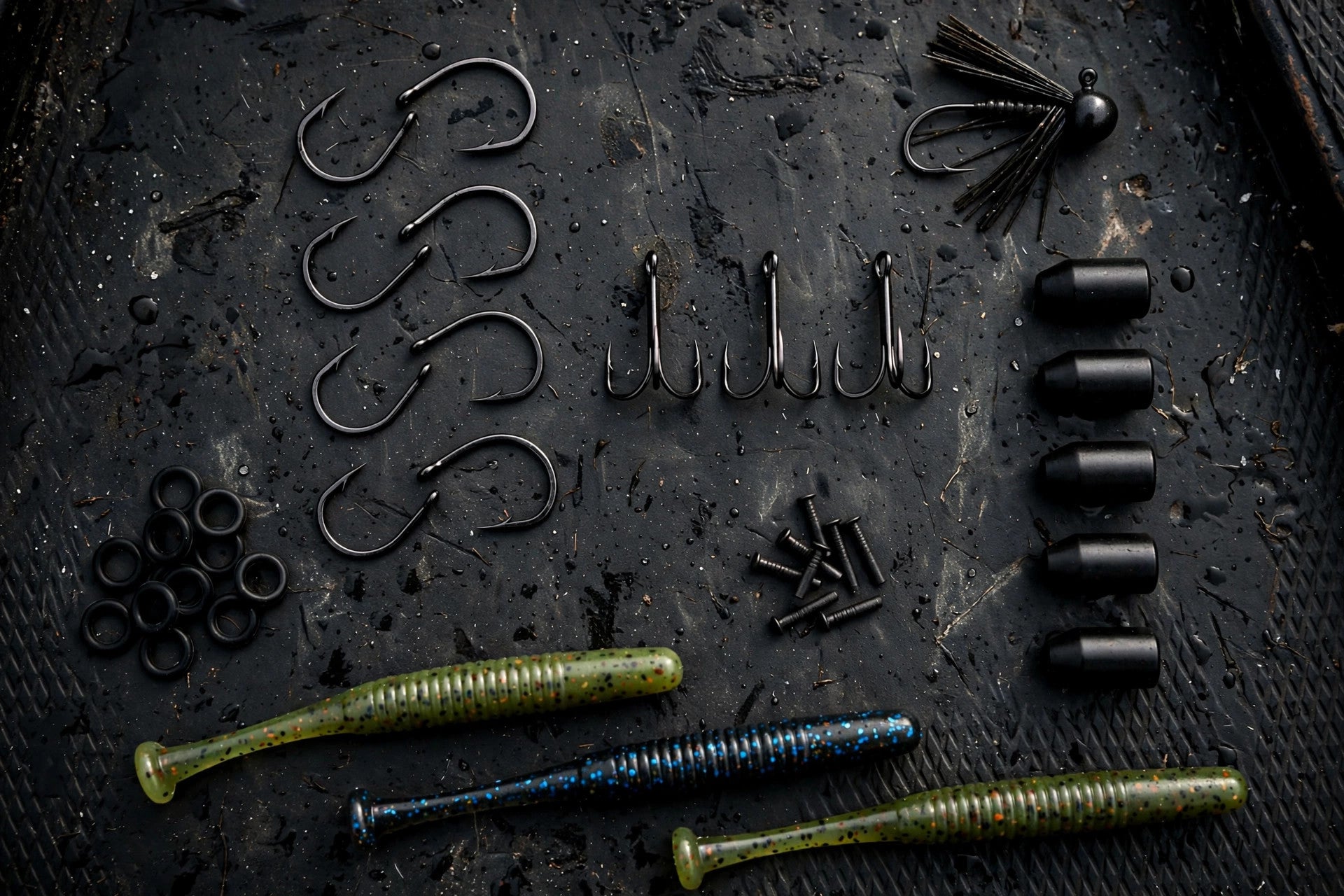 Flat-lay of bass fishing hooks, tungsten weights, O-rings and soft-plastic stick worms on a dark boat deck